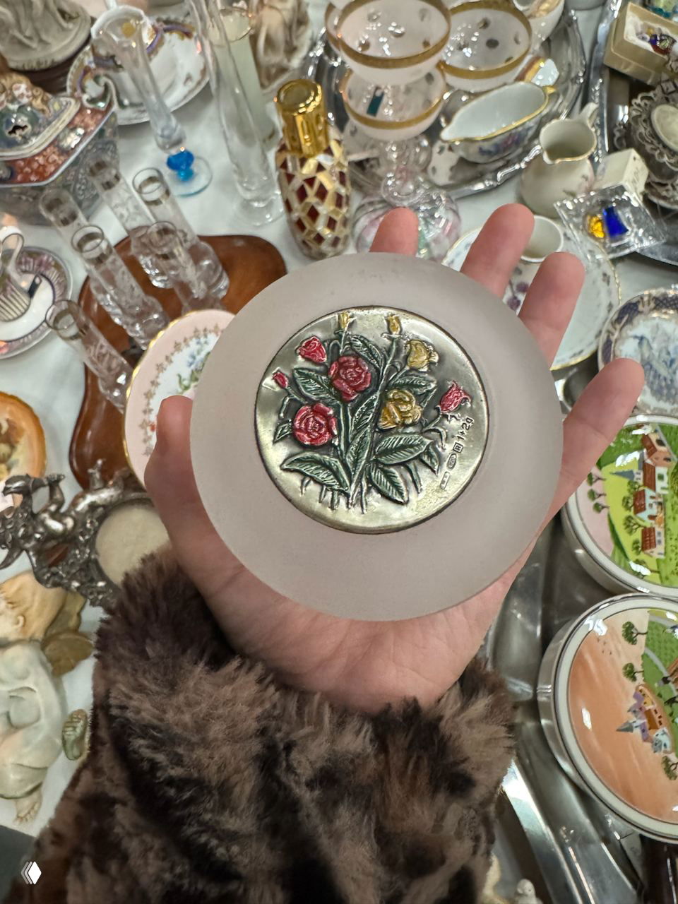 Ладонь держит небольшой декоративный сувенир с цветочным медальоном на фоне стола с множеством других мелких антикварных предметов.