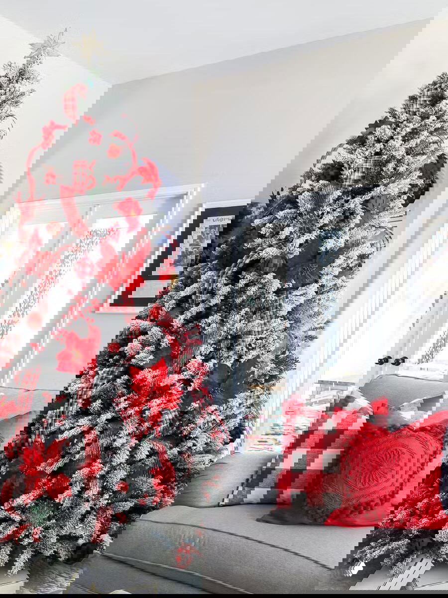 Пышная ель с красными акцентами, клетчатыми лентами и снежным эффектом, оформленная в традиционной красно‑белой гамме рядом с диваном и декоративными подушками.