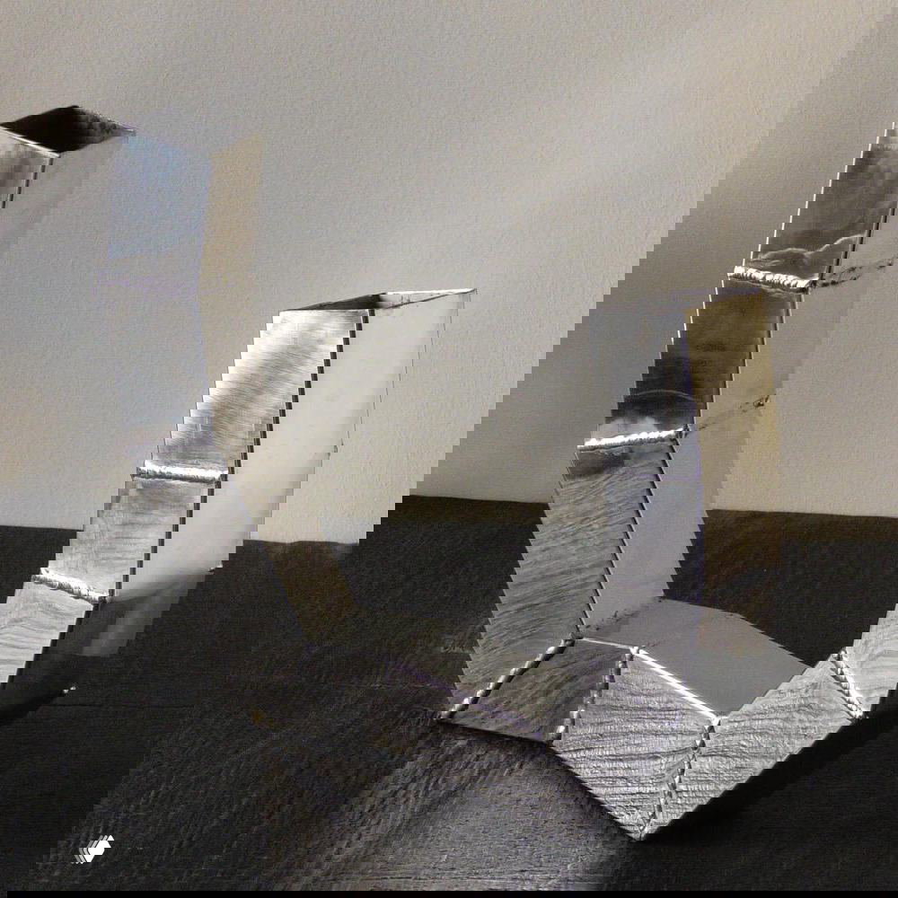 Геометрическая ваза или декоративный объект из сварных стальных элементов с видимыми швами и индустриальной фактурой на темной поверхности.