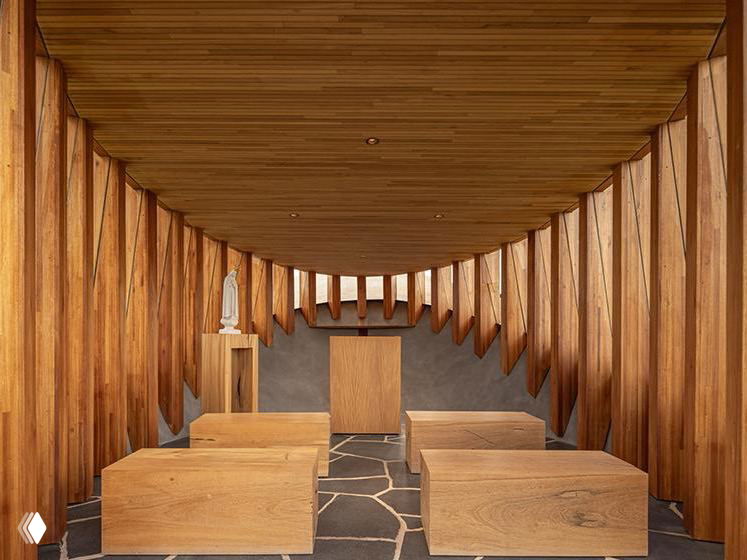 Интерьер капеллы: деревянные скамьи, алтарь, деревянные вертикальные ламели и каменный пол — минималистичное светлое пространство для молитвы и размышлений.