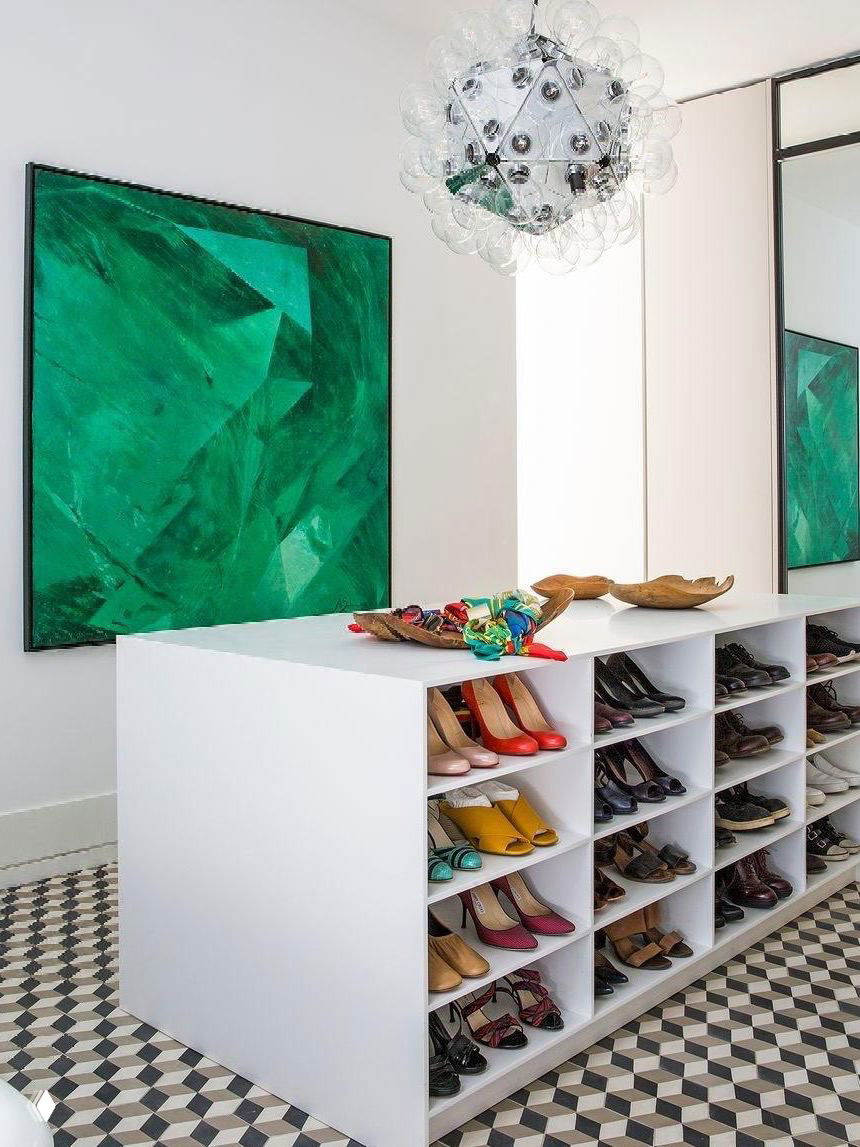 Островной белый комод с множеством квадратных отделений для обуви в центре комнаты; яркие туфли на полках и декоративная картина на стене.