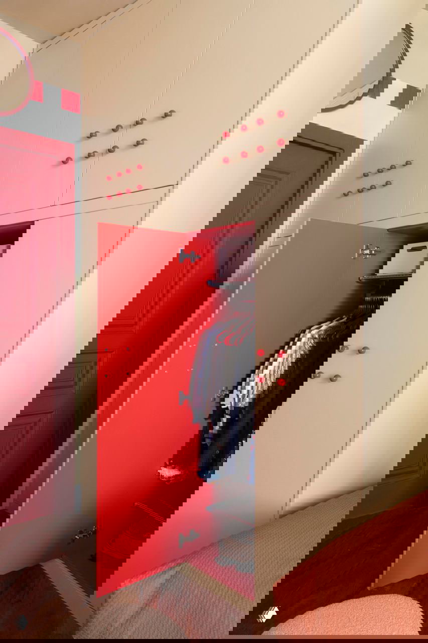 Встроенный шкаф бежевого цвета с ярко‑красной внутренней поверхностью двери, на которой видна одежда — практичное решение для хранения в детской.