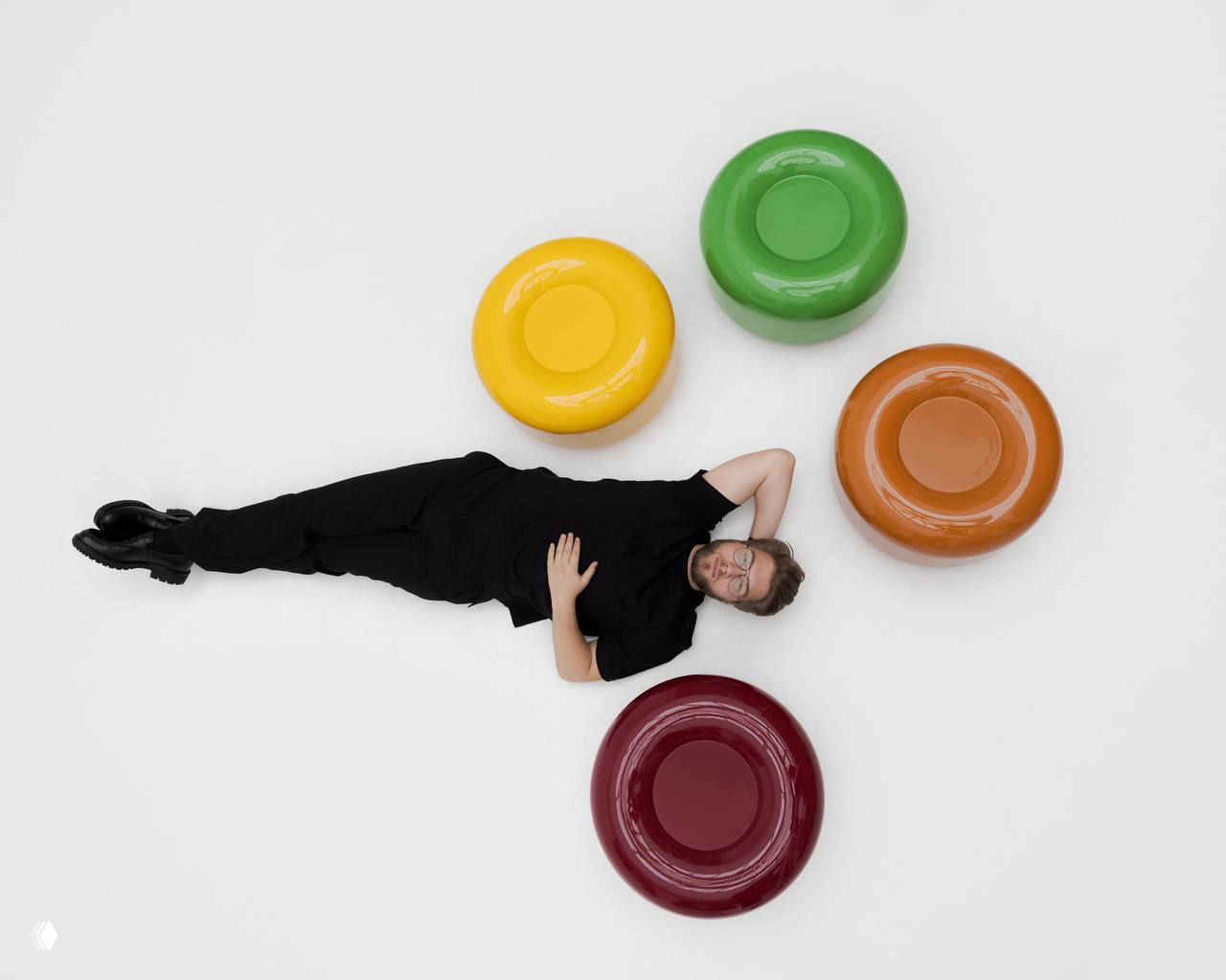 Дизайнер лежит на светлом полу рядом с круглыми пуфами-столиками Caramel в жёлтом, зелёном, коричневом и бордовом цветах, демонстрация масштаба предметов.