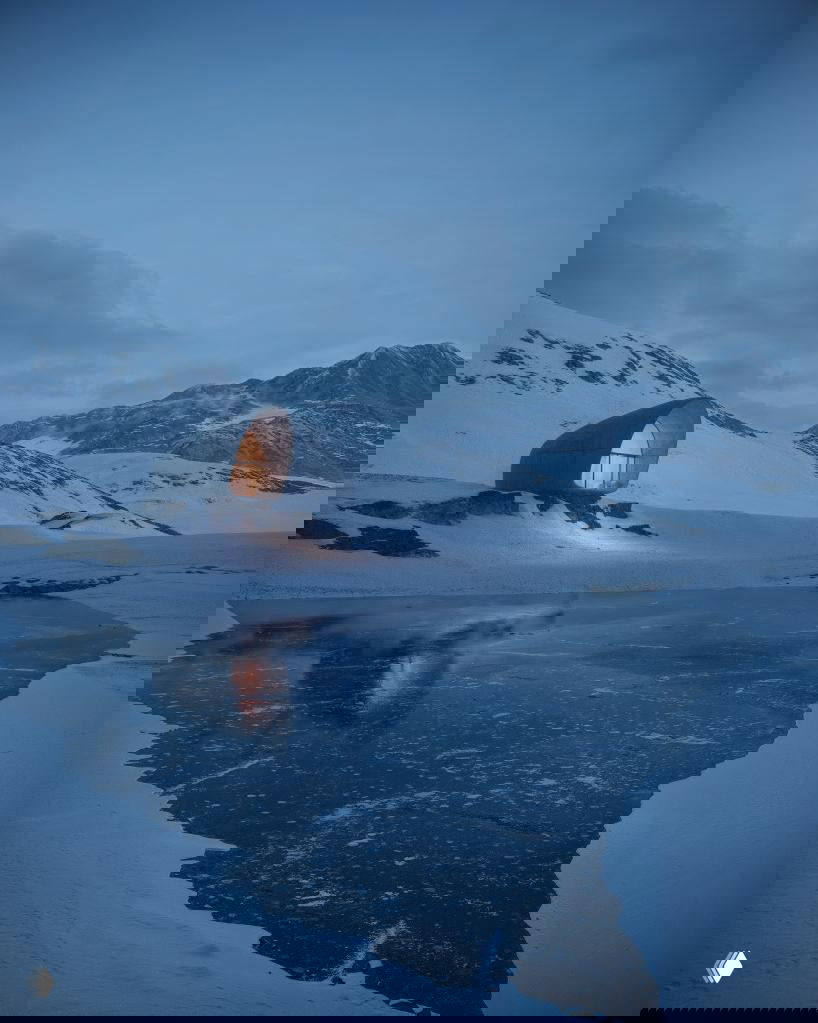 AltiHut у замёрзшего озера при сумерках: отражение подсвеченного фасада в ледяной воде и широкие заснеженные горы вокруг.