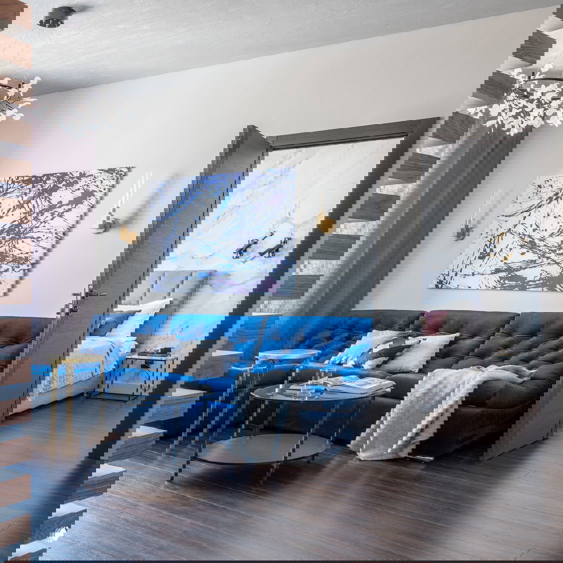 Современная гостиная с ярко-синим диваном и золотыми бра по бокам картины — сочетание насыщенного текстиля и точечного настенного освещения в комнате.