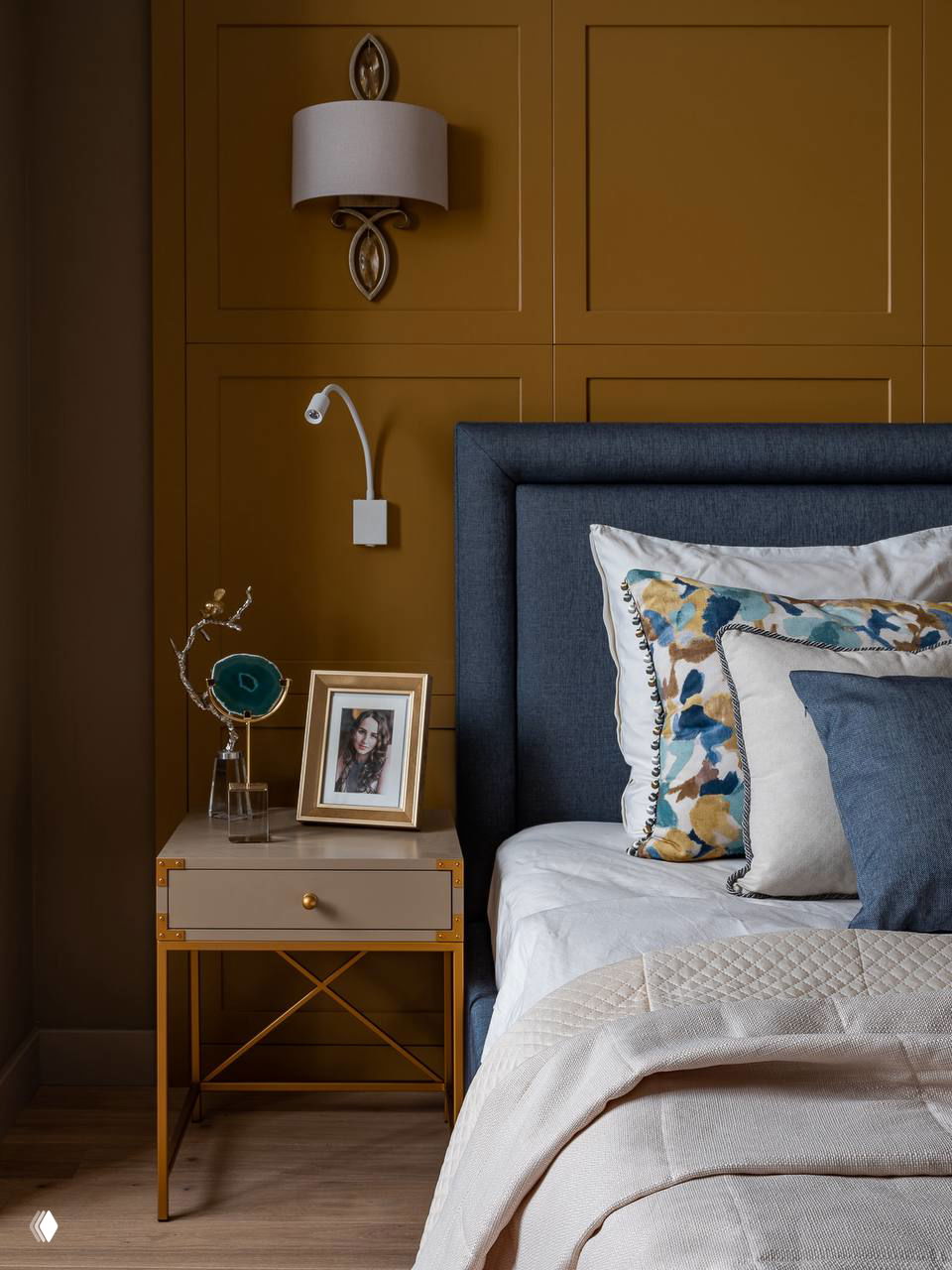 Акцентная оранжево‑коричневая стена за изголовьем, синее мягкое изголовье и декоративные подушки с цветочным принтом — художественный ход в спальне.
