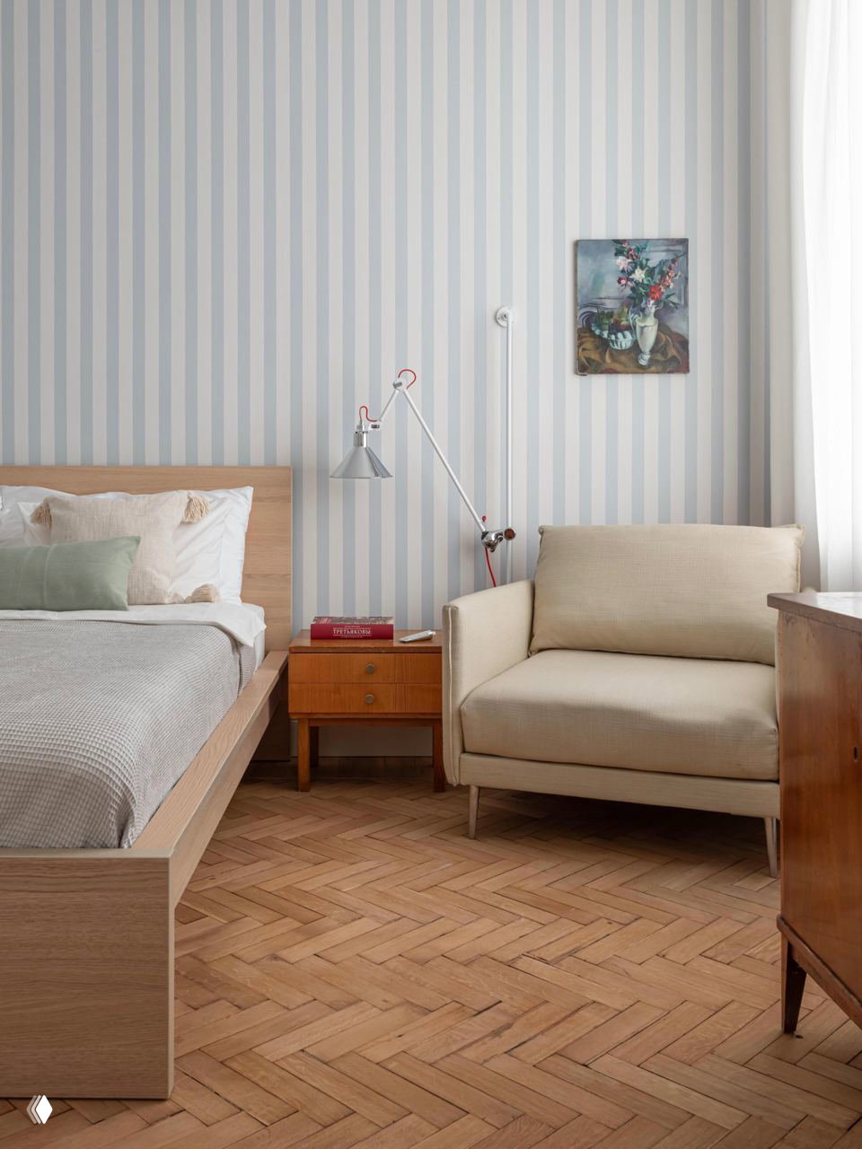 Светлая гостевая спальня с вертикальными полосатыми обоями, деревянной кроватью, тумбой и бежевым креслом — спокойный скандинавский интерьер для приёма гостей.