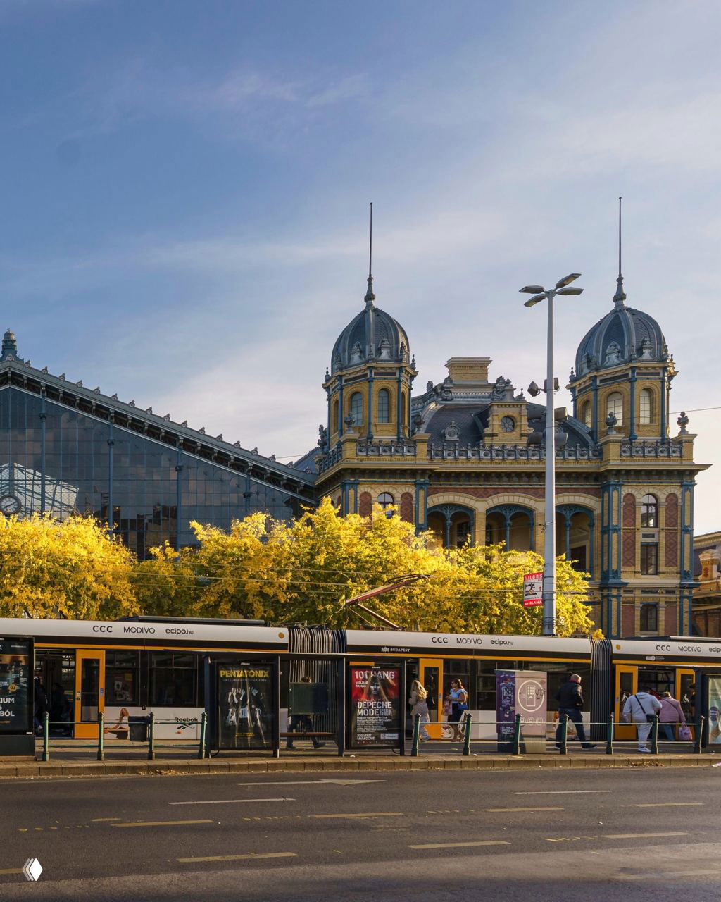 Городская улица с трамваем на переднем плане и архитектурным фасадом исторического вокзала или административного здания за ним, осенние деревья.
