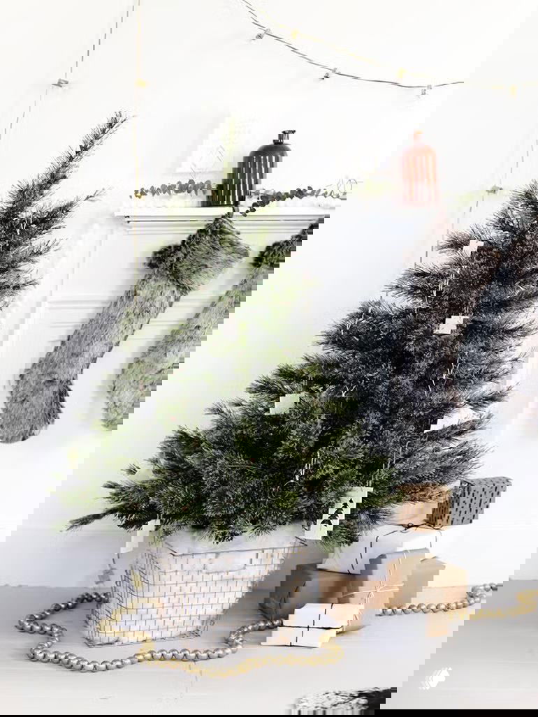 Небольшая ёлка в интерьерной композиции: корзина или кашпо, подарки вокруг и гирлянда из деревянных бусин у основания.