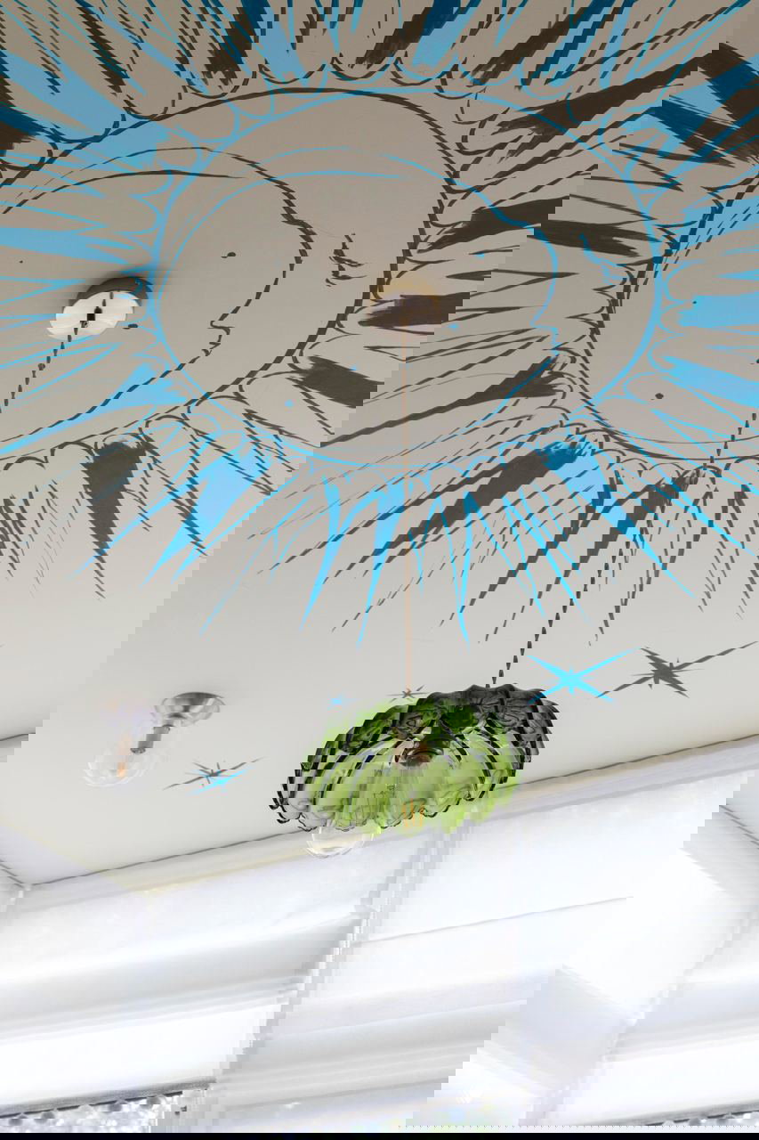 Фрагмент потолочного рисунка в виде солнечно‑лунного мотива синей краской и подвесной зелёный стеклянный плафон — художественный акцент интерьера.