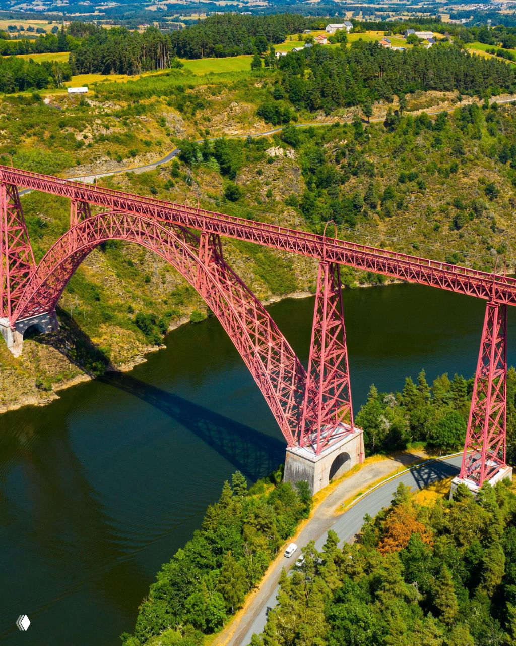 Красный металлический арочный мост через реку в окружении зелёных холмов: вид сверху на изогнутую конструкцию моста и водную поверхность.