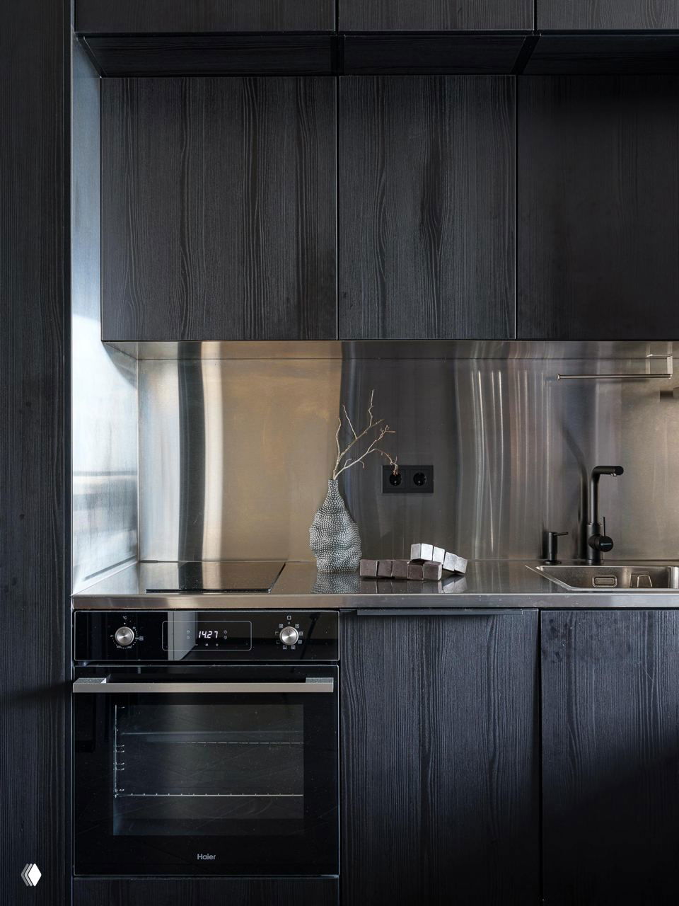 Крупный план кухонной рабочей зоны: индукционная плита, металлический фартук и минималистичная мойка на фоне тёмных деревянных фасадов.