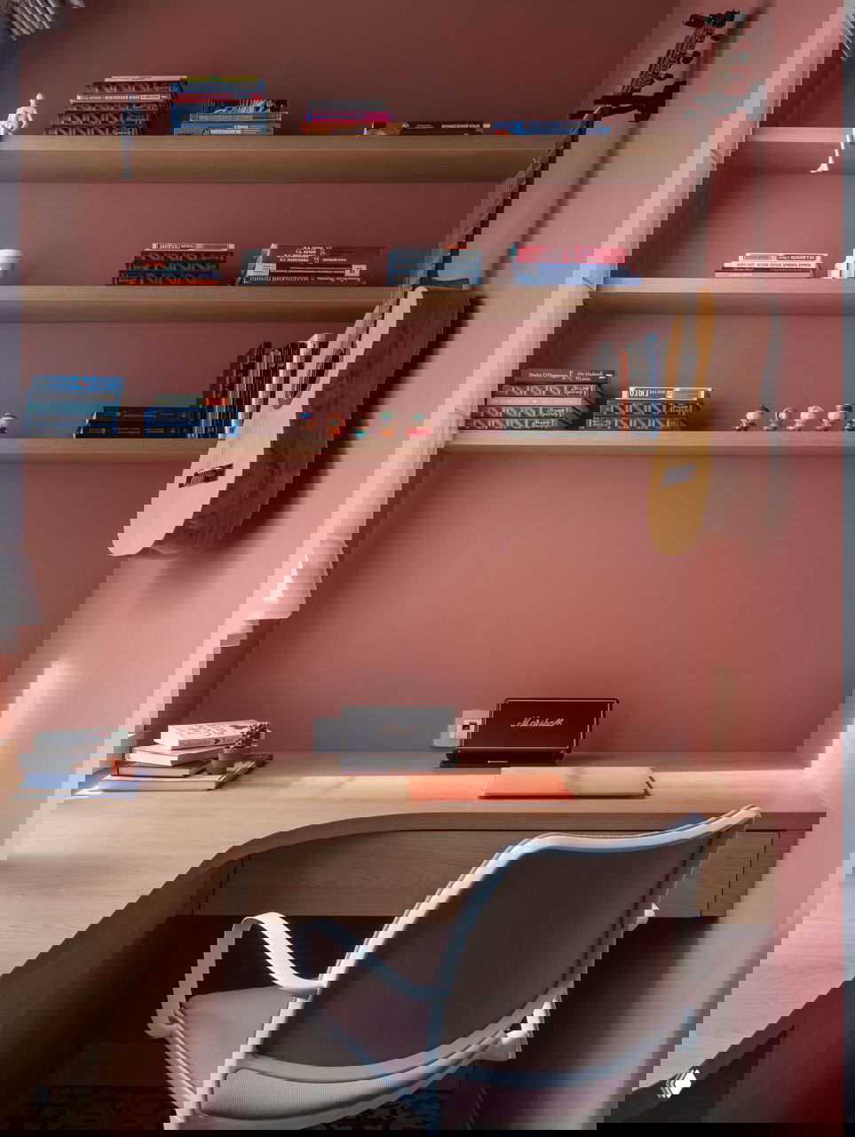 Рабочая зона с пастельно-розовой стеной, встроенными деревянными полками и висящей гитарой; компактный стол и стул демонстрируют, как покраска стены и полки могут изменить комнату.