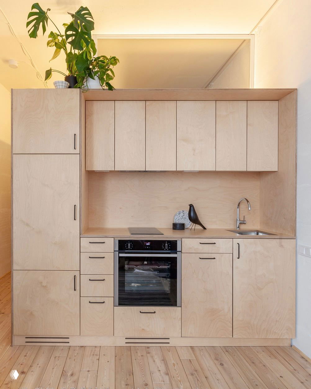 Минималистичная кухня с аккуратным пеналом и встроенной духовкой: фасады из светлой фанеры, небольшой декоративный букет и чистая линейная композиция рабочей зоны.