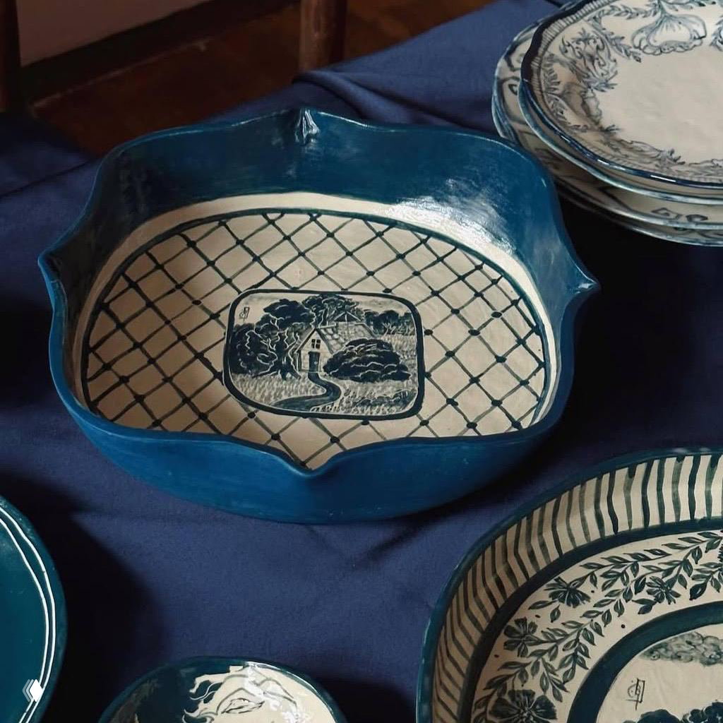 Глубокая керамическая чаша тёмно‑бирюзового цвета с центральным изображением и сетчатым орнаментом, выставленная на столе вместе с тарелками — авторская посуда.