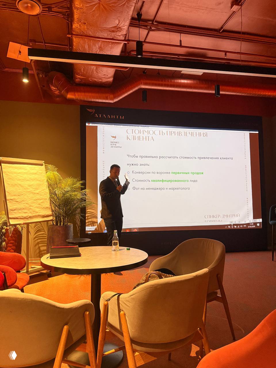 Спикер у большого экрана с маркерами и текстом по расчёту стоимости привлечения клиента, вокруг кресла и столы — вид зала и сцены
