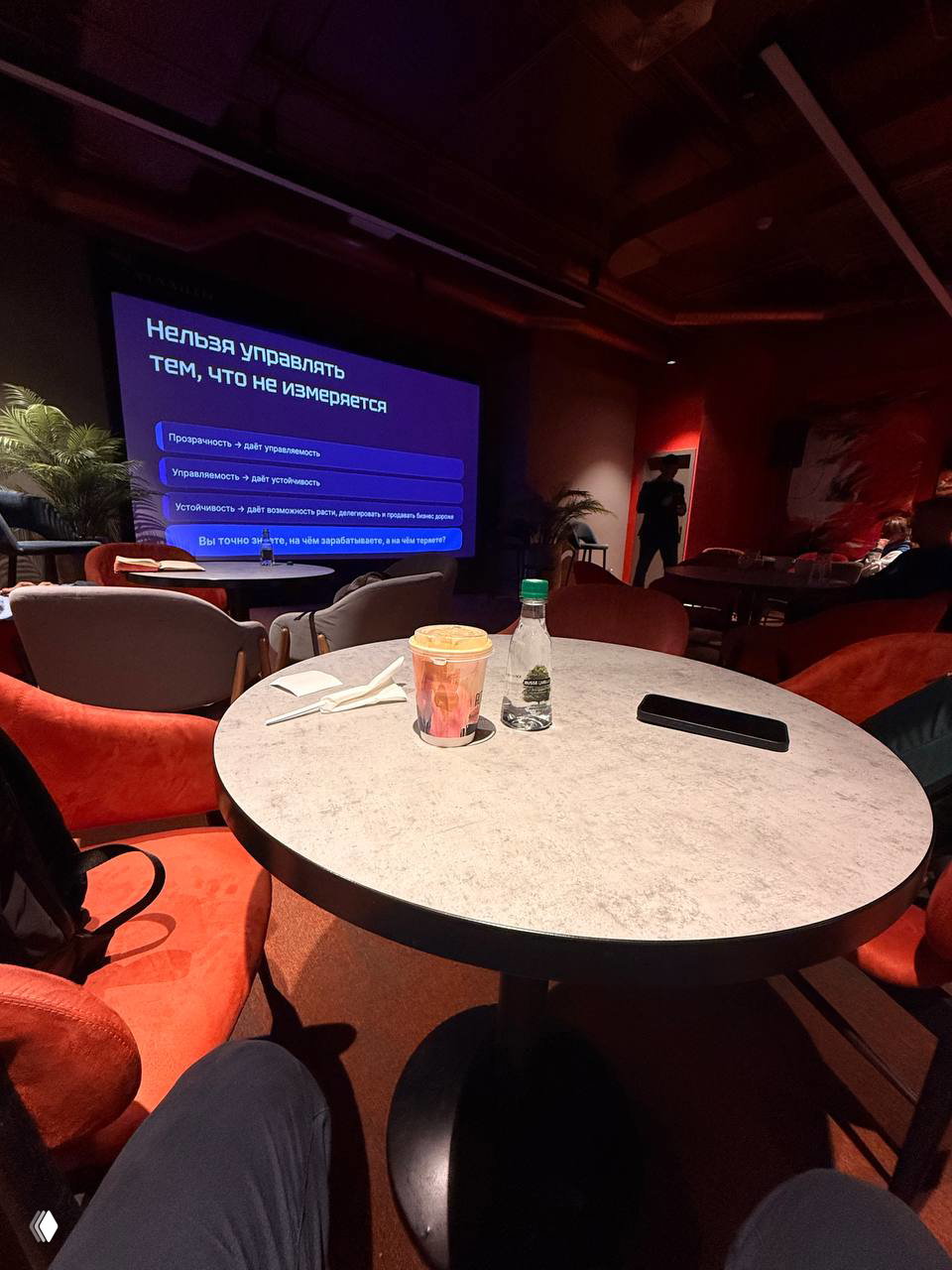 Круглый стол в аудитории, на столе стакан кофе и бутылка воды, вдалеке экран с текстом «Нельзя управлять тем, что не измеряется» — общая сцена мероприятия