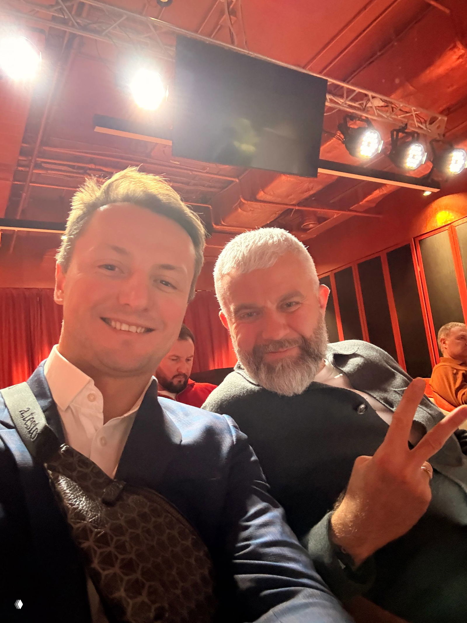 Селфи двух мужчин в аудитории с тёплым освещением; один показывает жест «V», сцена и зрители видны на заднем плане во время мастер‑класса.