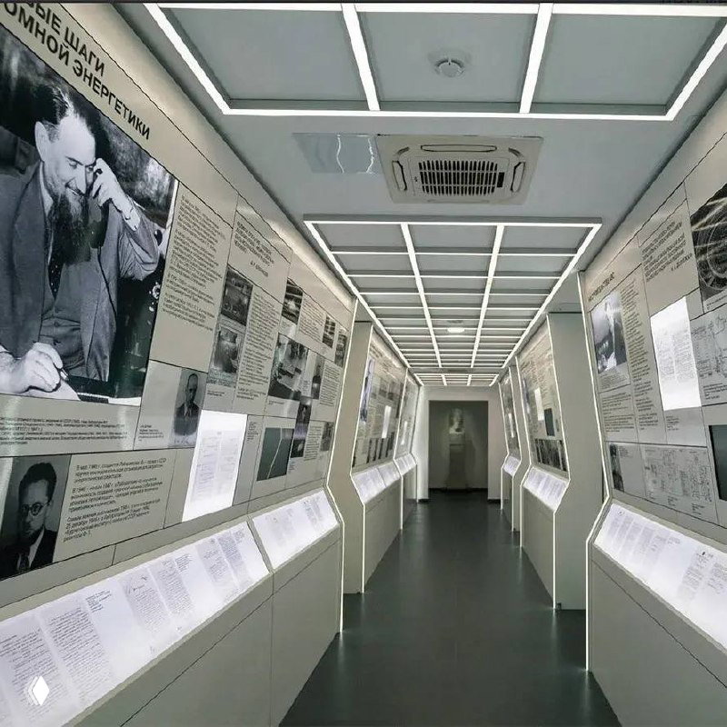 Коридор музейной экспозиции с интегрированными потолочными LED-панелями и подсветкой витрин; нейтральный ровный свет обеспечивает читаемость материалов и вывесок.