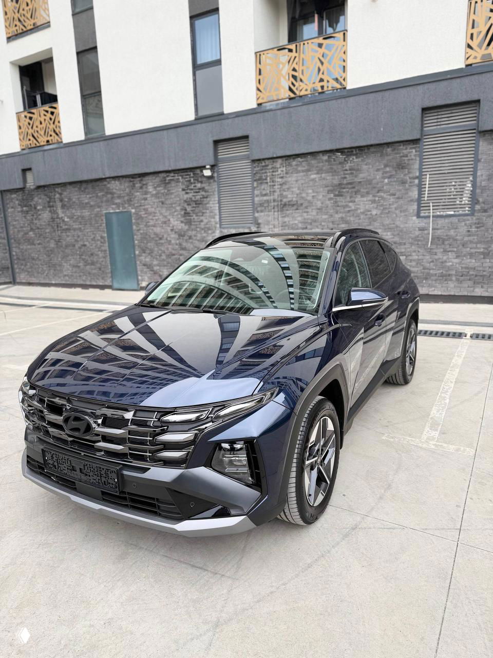 Тёмно‑синий Hyundai Tucson 2025 на стоянке под углом; видны R18 диски, отражения зданий на капоте и общие пропорции кузова.