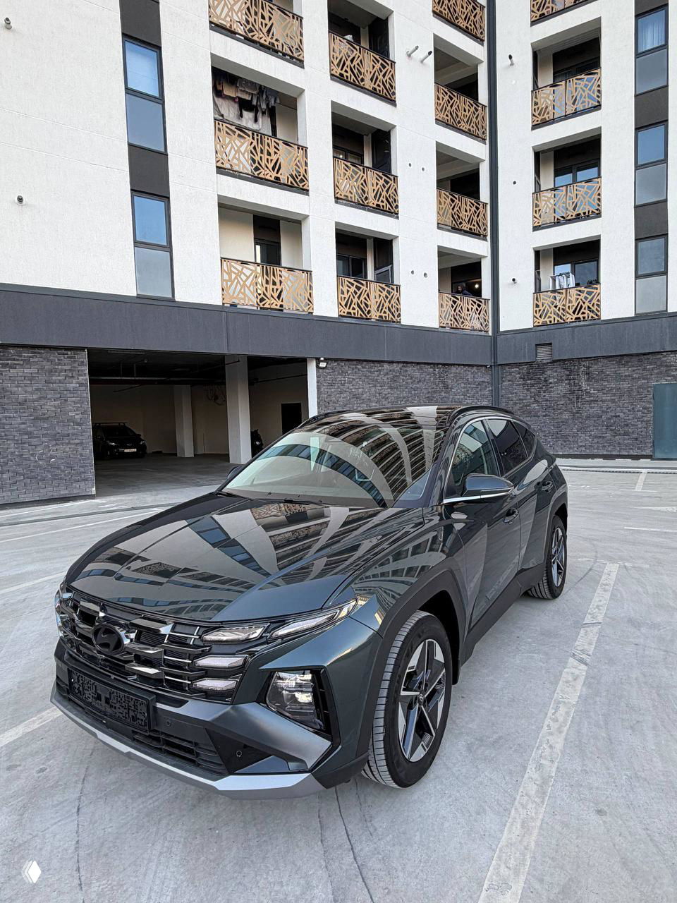 Тёмно‑серый Hyundai Tucson 2025, общий вид сбоку на фоне жилого комплекса, видны линии кузова, крыша и профиль автомобиля.