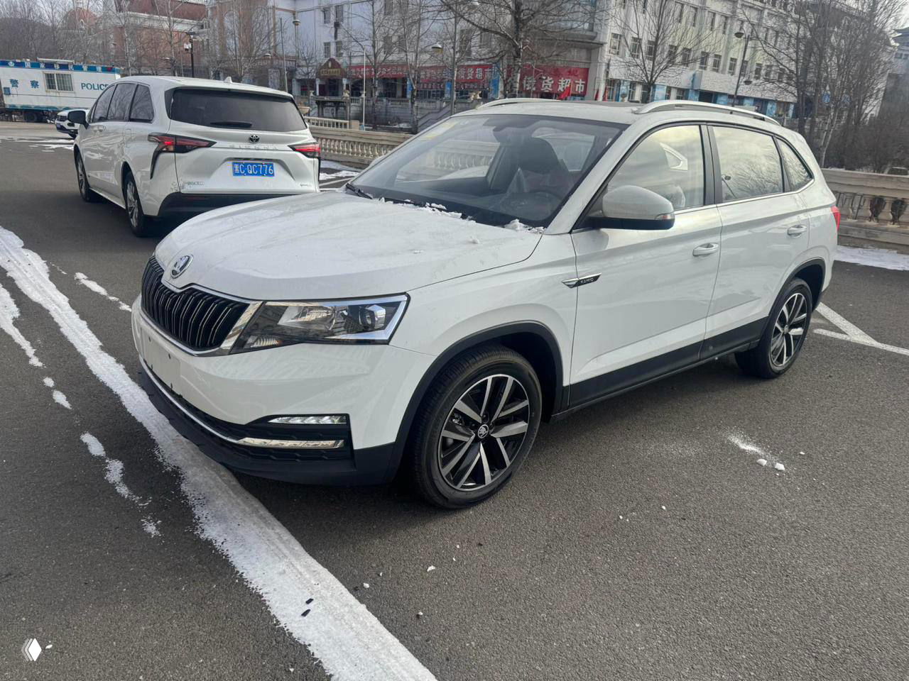 Боковой общий план белой Skoda Kamiq на стоянке в Китае, автомобиль на асфальте с небольшими следами снега и окрестными машинами.