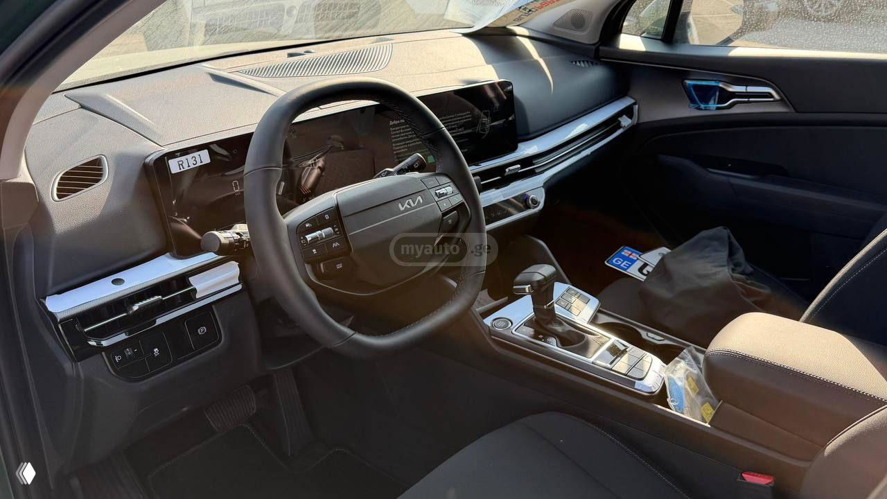Салон KIA Sportage 2025, общий вид приборной панели и мультимедийного дисплея, руль с логотипом KIA и центральная консоль в дневном свете.