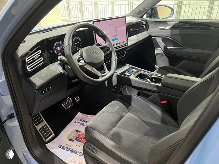 Фронтальный вид салона Volkswagen Tiguan L Pro: кресла, центральный тоннель и элементы управления, отделка в тёмных тонах.