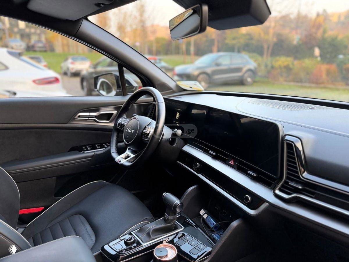 Интерьер KIA Sportage: руль с логотипом KIA, большая панель приборов и сенсорный экран, центральная консоль и селектор АКПП на переднем плане.
