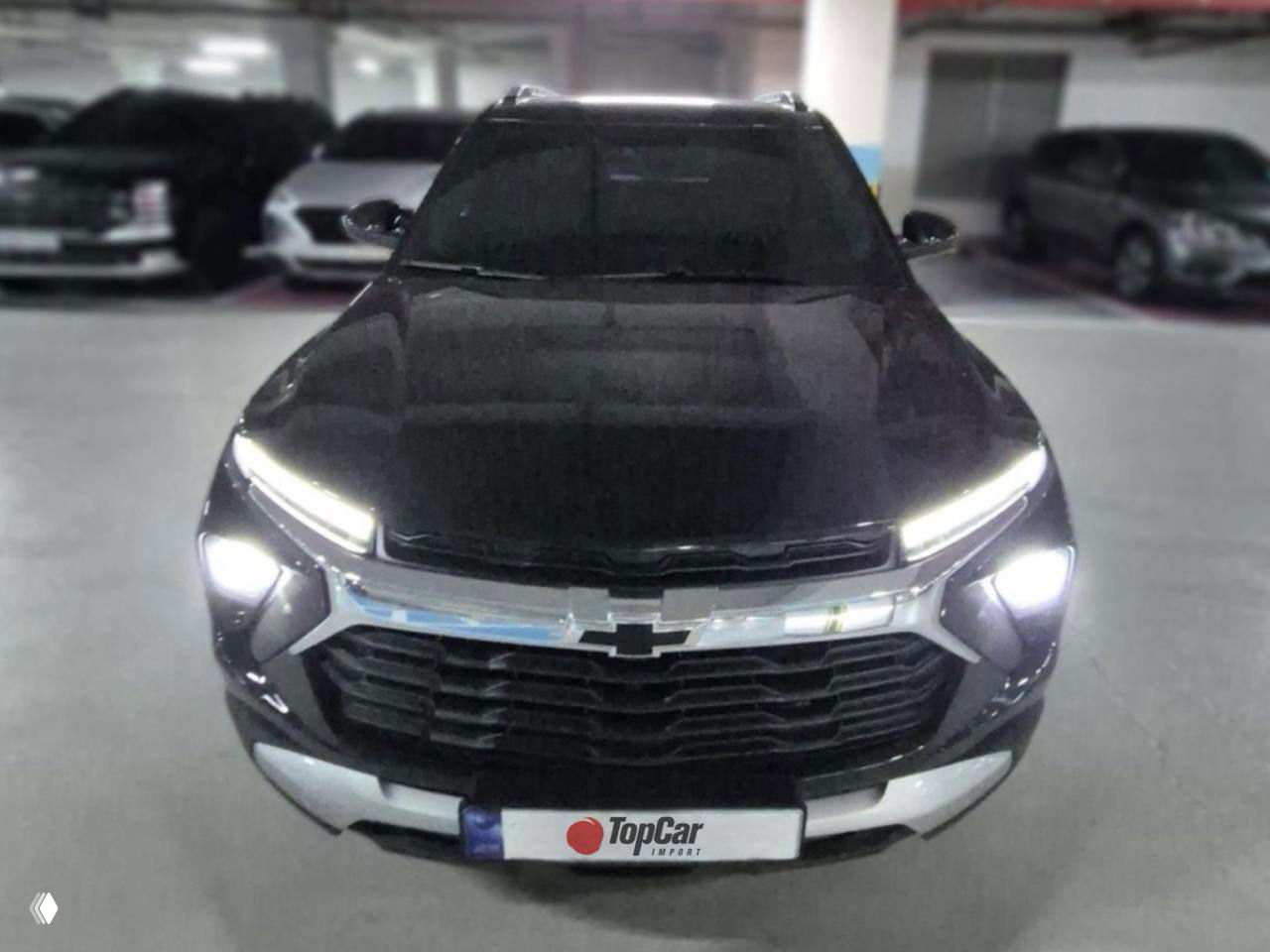 Чёрный Chevrolet Trailblazer в помещении, включённые фары и фирменная номерная пластина TopCar, фотография подчёркивает переднюю оптику и решётку.