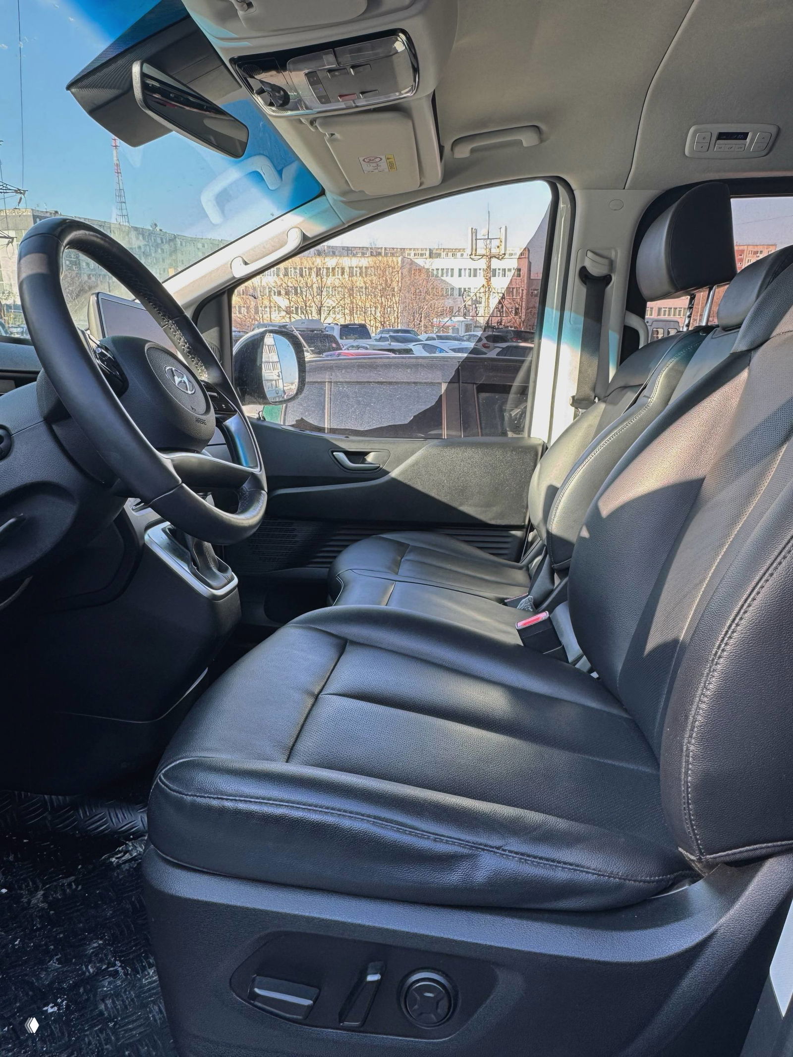 Вид на салон Hyundai Staria — передние кожаные сиденья и руль, состояние обивки и вид через боковое окно на городской паркинг.