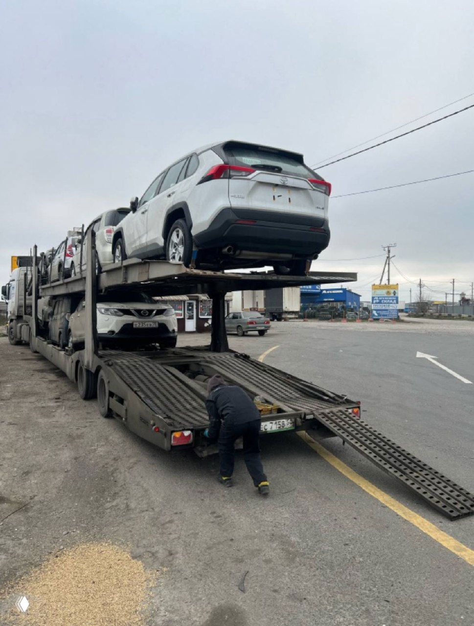 Рабочие готовят аппарель автовоза для выгрузки, вид сзади на трейлер с автомобилями; уличная стоянка и дорожные указатели на заднем плане