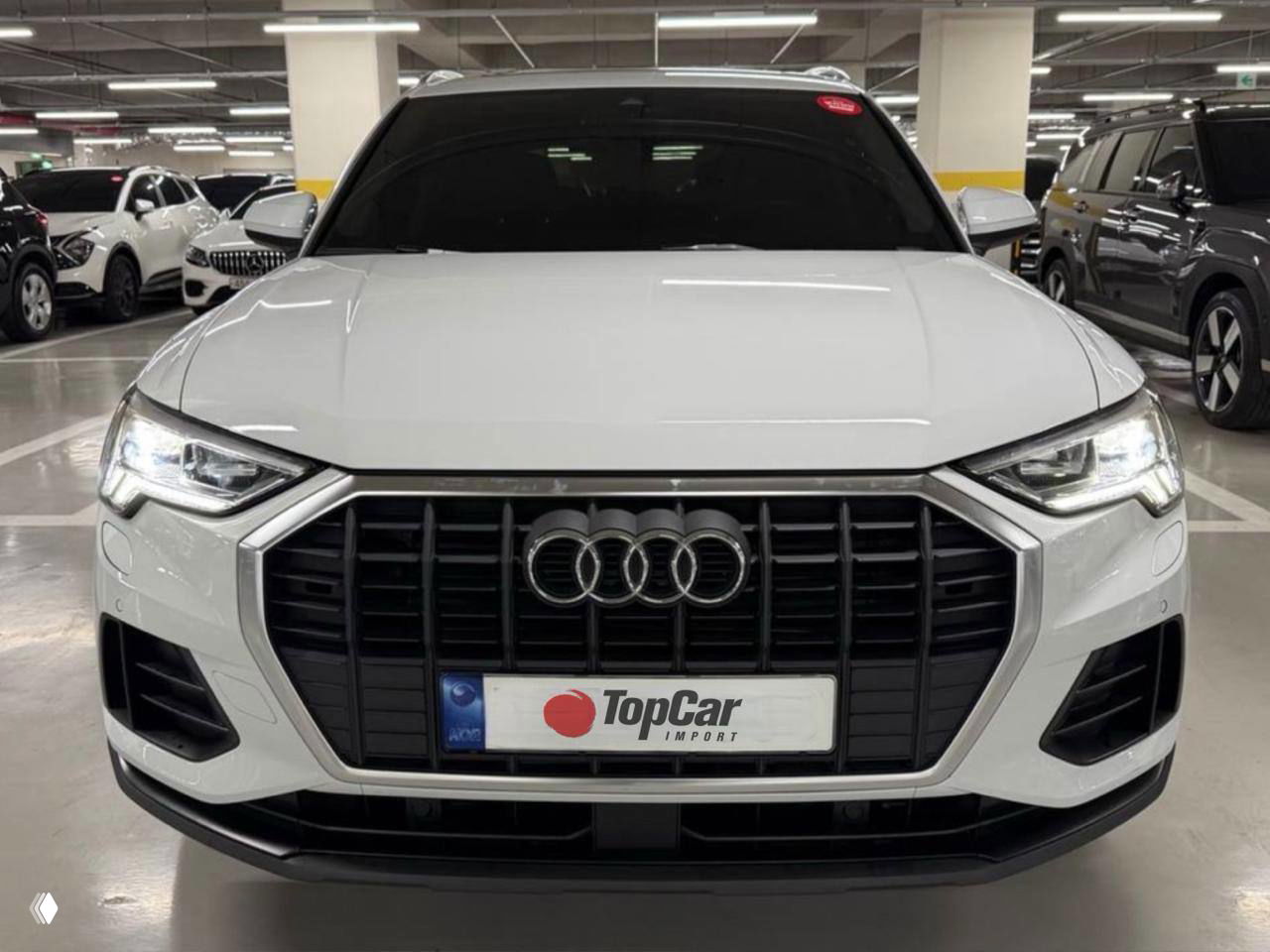 Передняя часть белого Audi Q3 в крытой парковке, чётко видна фирменная решётка Audi и номерная пластина с логотипом TopCar.
