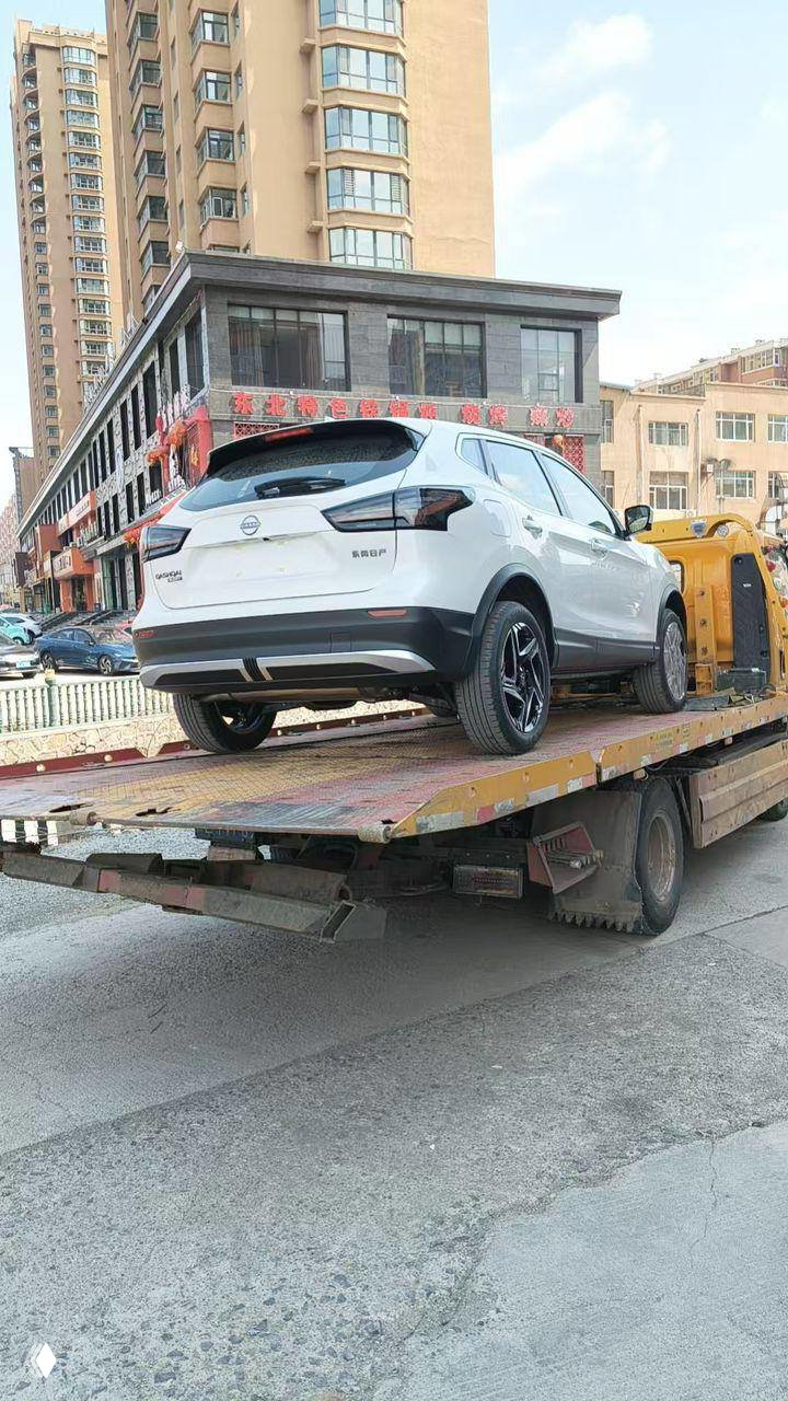 Белый Nissan Qashqai, вид сзади на платформе эвакуатора во время доставки; городской фон с многоэтажными зданиями на заднем плане.