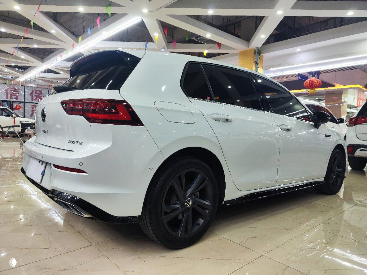 Вид сзади и сбоку белого Volkswagen Golf R‑Line в автосалоне, демонстрирующий форму задней части, фары и выхлопную отделку