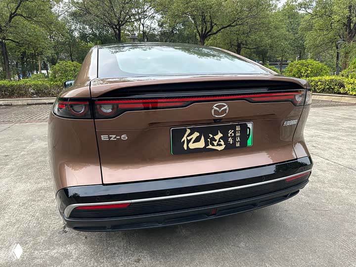 Задняя часть Mazda EZ-6 с шильдиком модели и номерной табличкой, демонстрация рисунка задних фонарей и линии багажника на фоне зелёных деревьев.