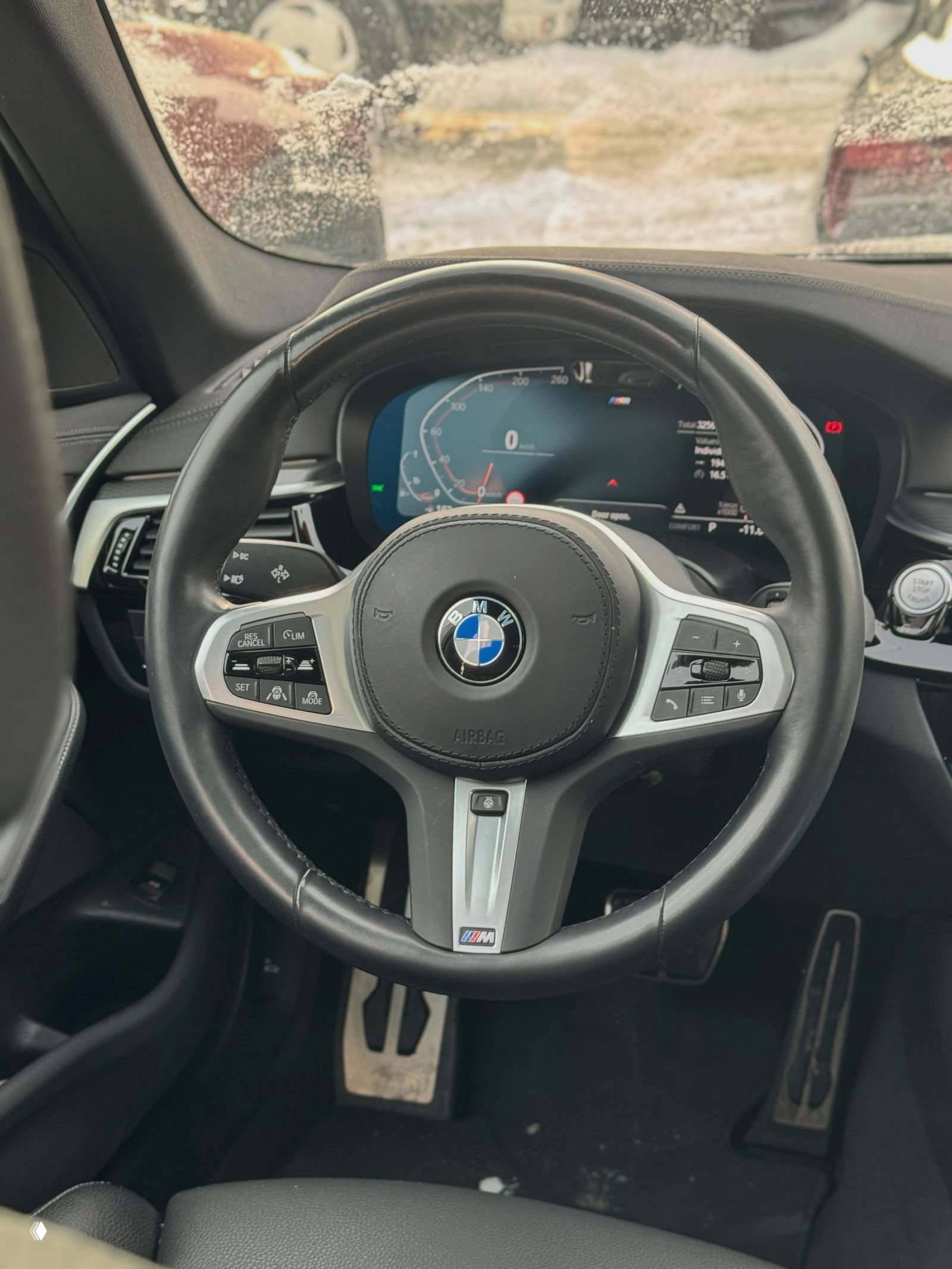 Интерьер BMW 520i: рулевое колесо с логотипом BMW и элементами M‑пакета, вид на цифровую приборную панель и элементы управления, частично покрытые снегом снаружи.