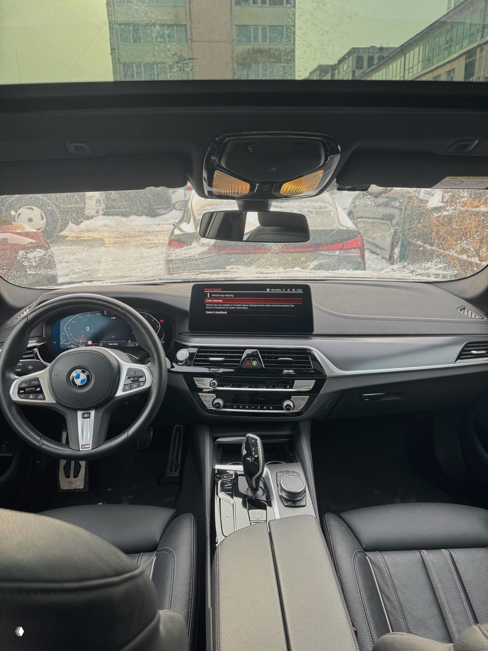 Общий вид салона BMW 520i 2022: передняя консоль, селектор АКПП и панорамное лобовое стекло с видом на стоянку, аккуратный кожаный интерьер.