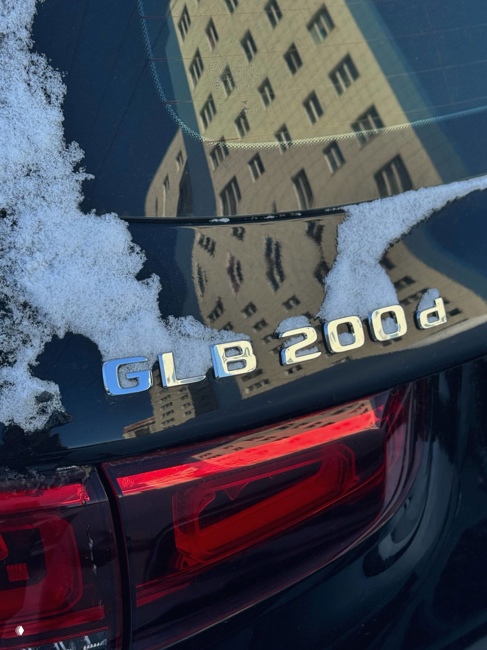 Крупный план эмблемы и шильдика GLB 200d на крышке багажника, частично покрытой снегом, с отражением окружающего здания на поверхности.