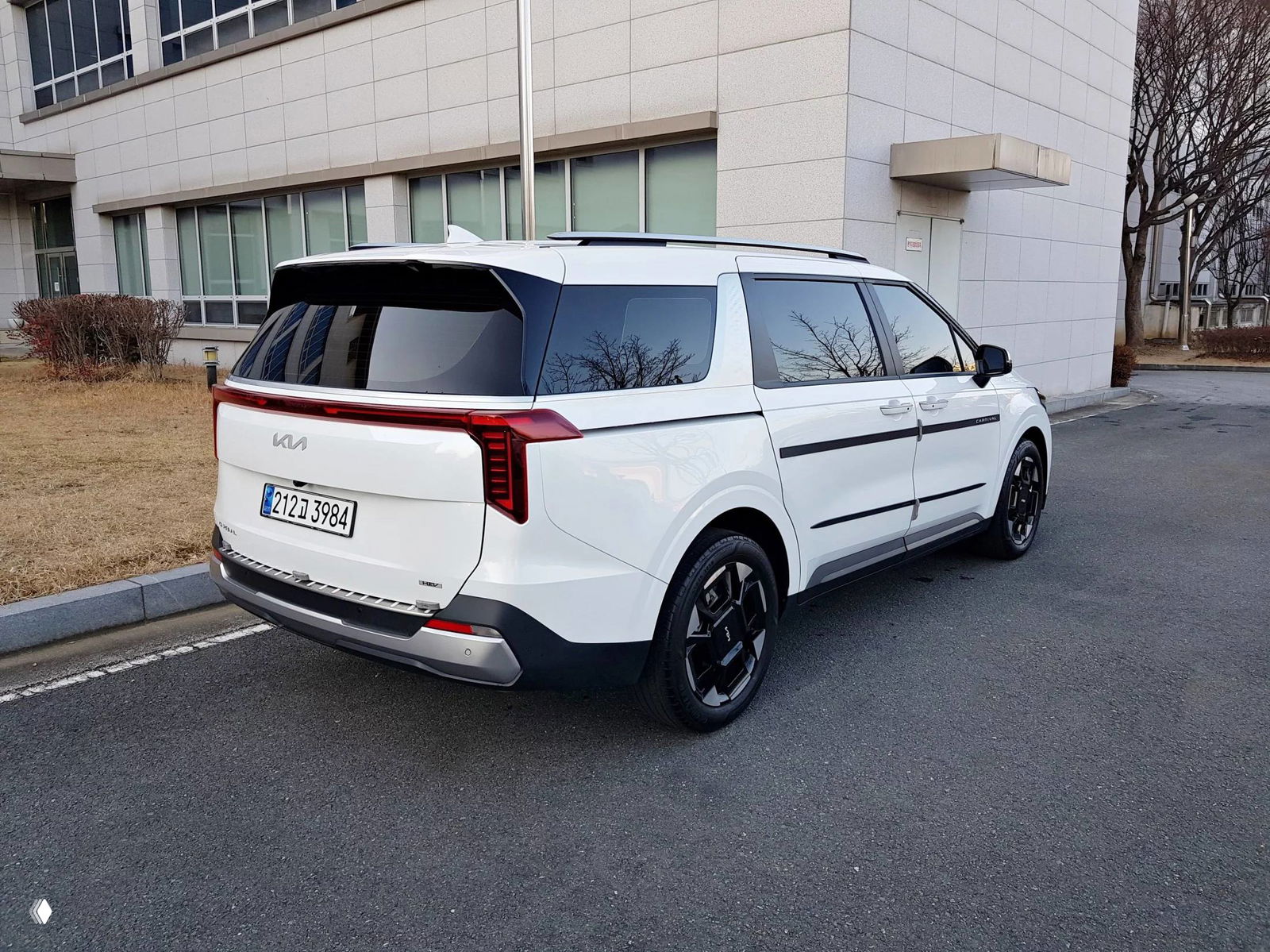 KIA Carnival белого цвета, вид сзади и сбоку на стоянке, видны задние фонари в виде тонкой световой полосы и задняя дверь минивэна.