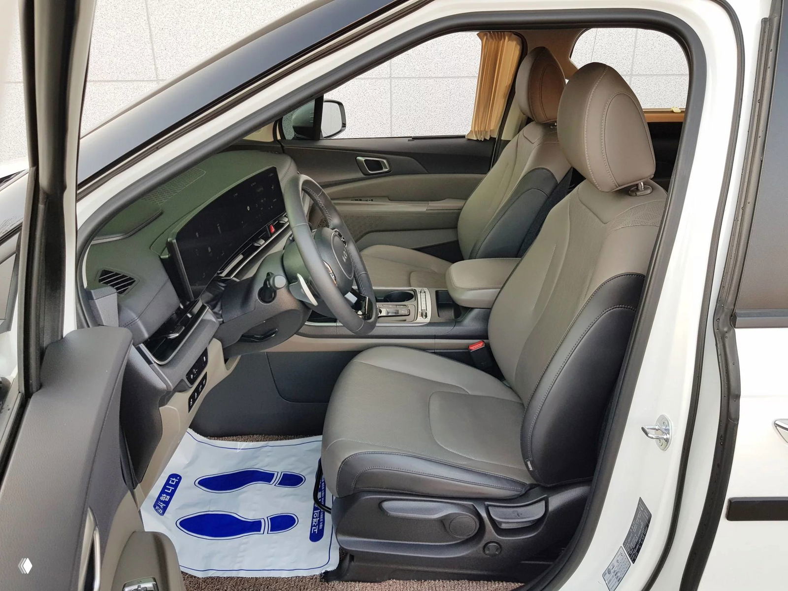 Вид на передние сиденья KIA Carnival: сиденье водителя в нейтральной коже, подлокотник и центральная консоль с регулятором трансмиссии.