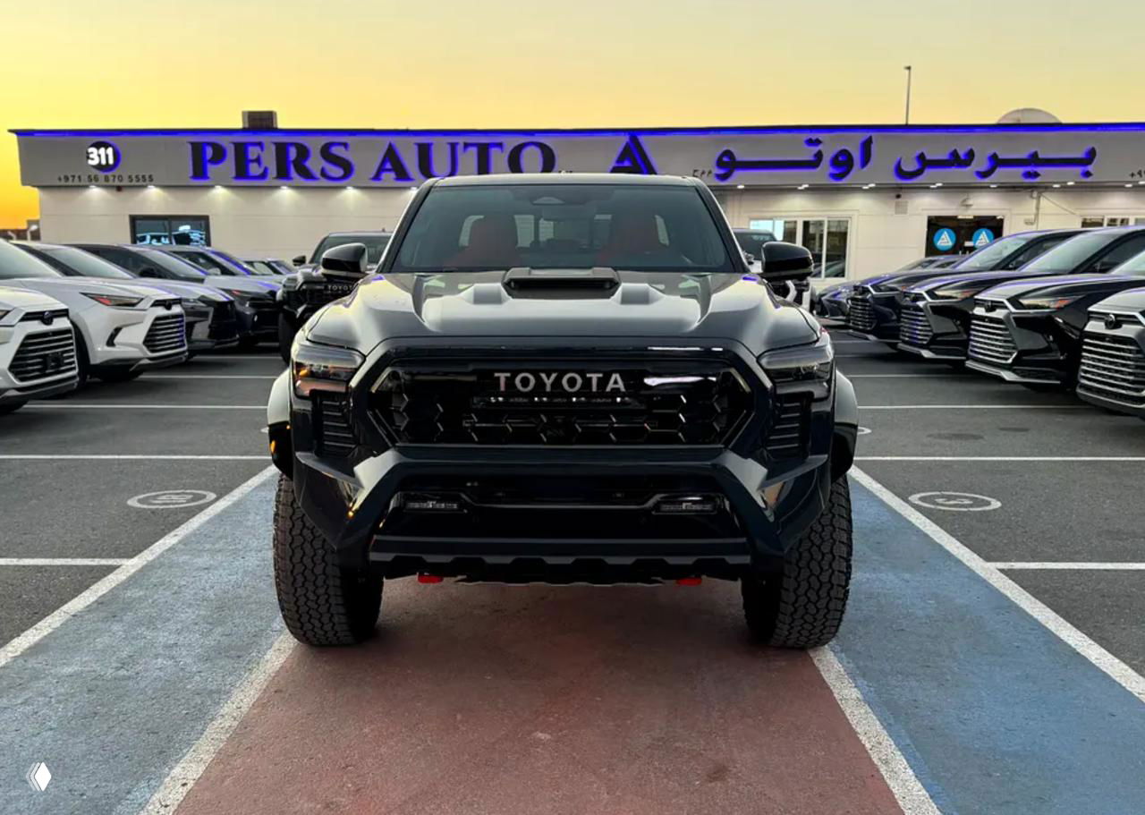 Фронтальный вид Toyota Tacoma TRD 2025 у дилерского центра; крупный план решётки радиатора с надписью TOYOTA и агрессивный бампер автомобиля.