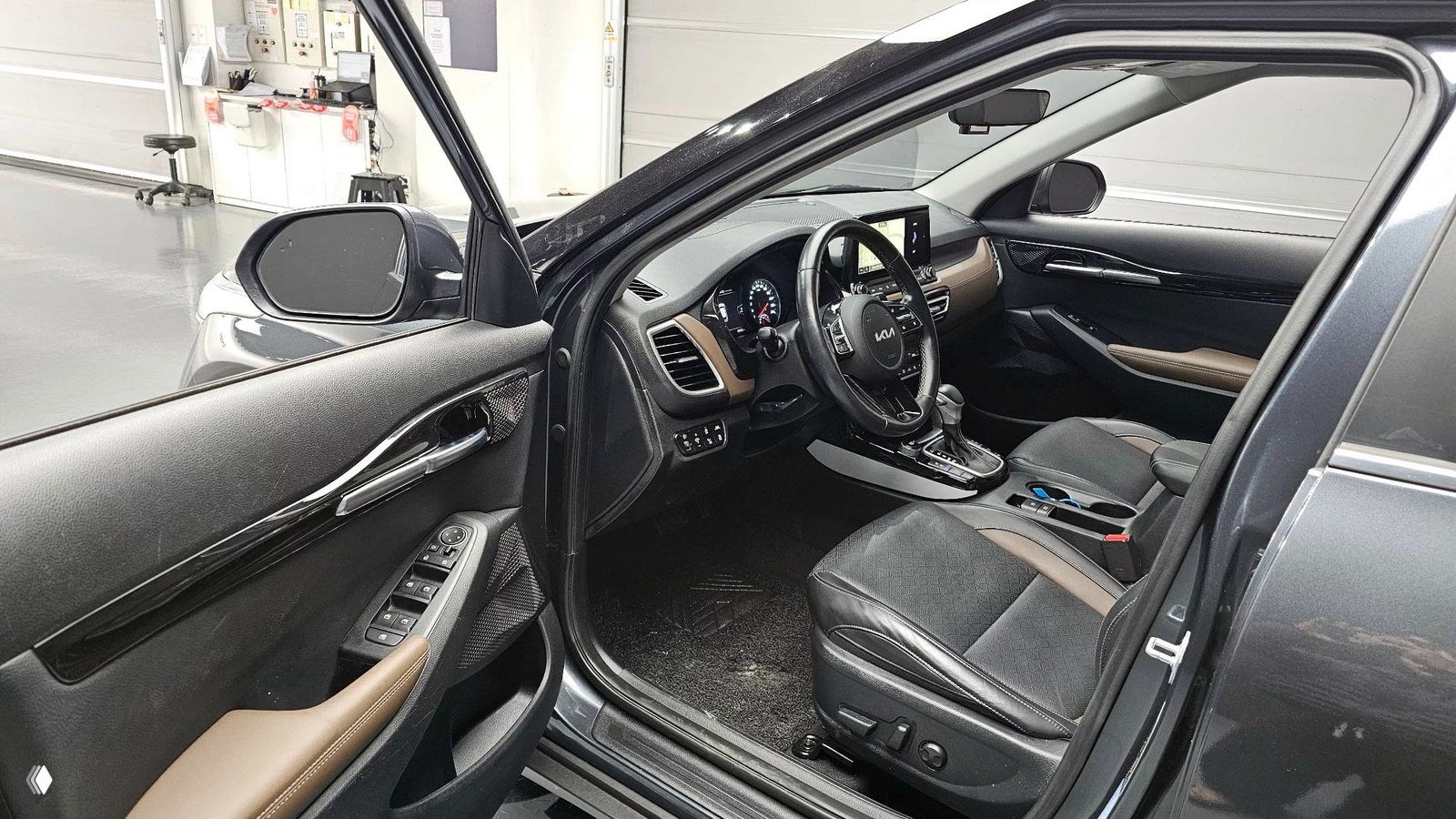 Открытая водительская дверь KIA Seltos: вид на переднее сиденье, руль с логотипом KIA и элементы управления на дверной панели, общая перспектива салона.