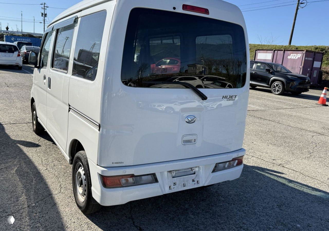 Вид сзади белого Daihatsu HIJET S321V на аукционной площадке, видно заднюю дверь, эмблему HIJET и общей фон стоянки.