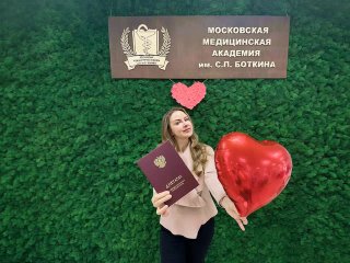Девушка улыбается, держит диплом ММА и красный шар в форме сердца на зелёной стене с эмблемой и табличкой академии