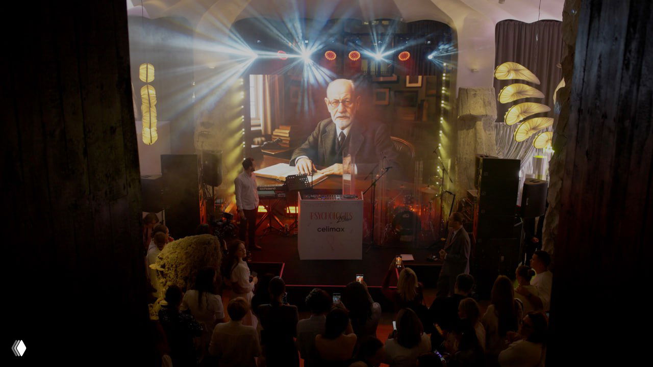 Вид сверху на сцену с прожекторами и большим экраном, на котором проецируют исторический образ; гости внизу наблюдают за программой вечера.