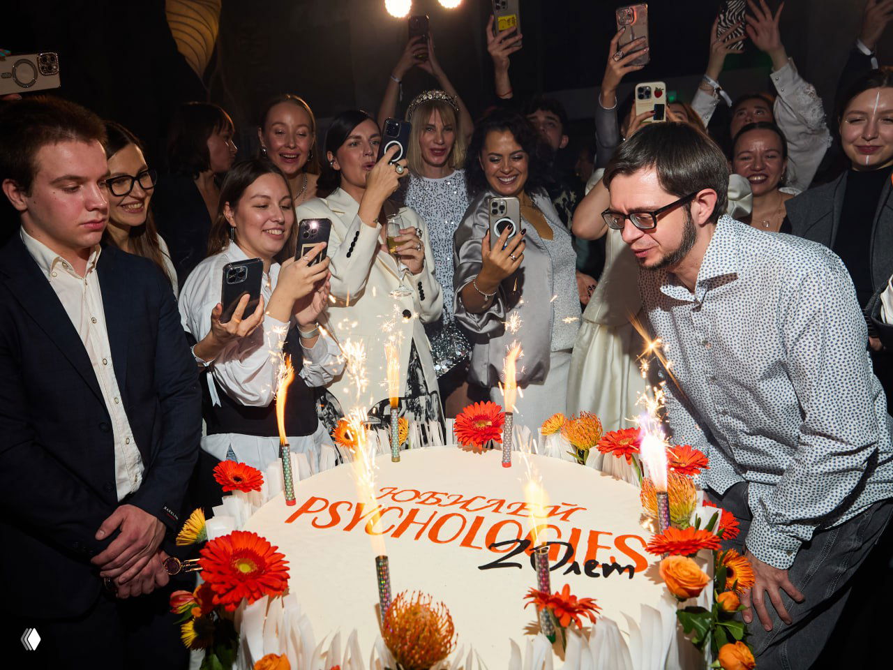 Праздничный торт с надписью в честь юбилея PSYCHOLOGIES окружён гостями, многие снимают момент на смартфоны, искры и свечи создают праздничную сцену.