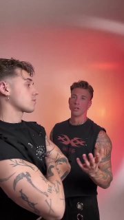 Два молодого человека с татуировками обсуждают что-то в студии с тёплой подсветкой; кадр показывает взаимодействие авторов контента.