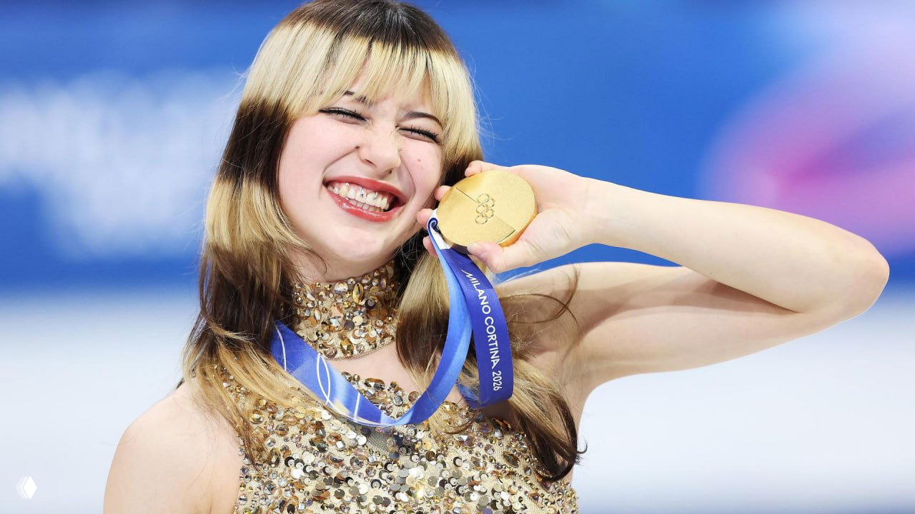 Крупный план улыбающейся 20‑летней фигуристки Алисы Лью, держащей золотую медаль Milano Cortina 2026 на синем фоне льда и трибуны.