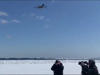 БПЛА (дрон) летит над заснеженным морским побережьем Сахалина; рядом операторы и фотографы в зимней одежде фиксируют и наблюдают за полётом.
